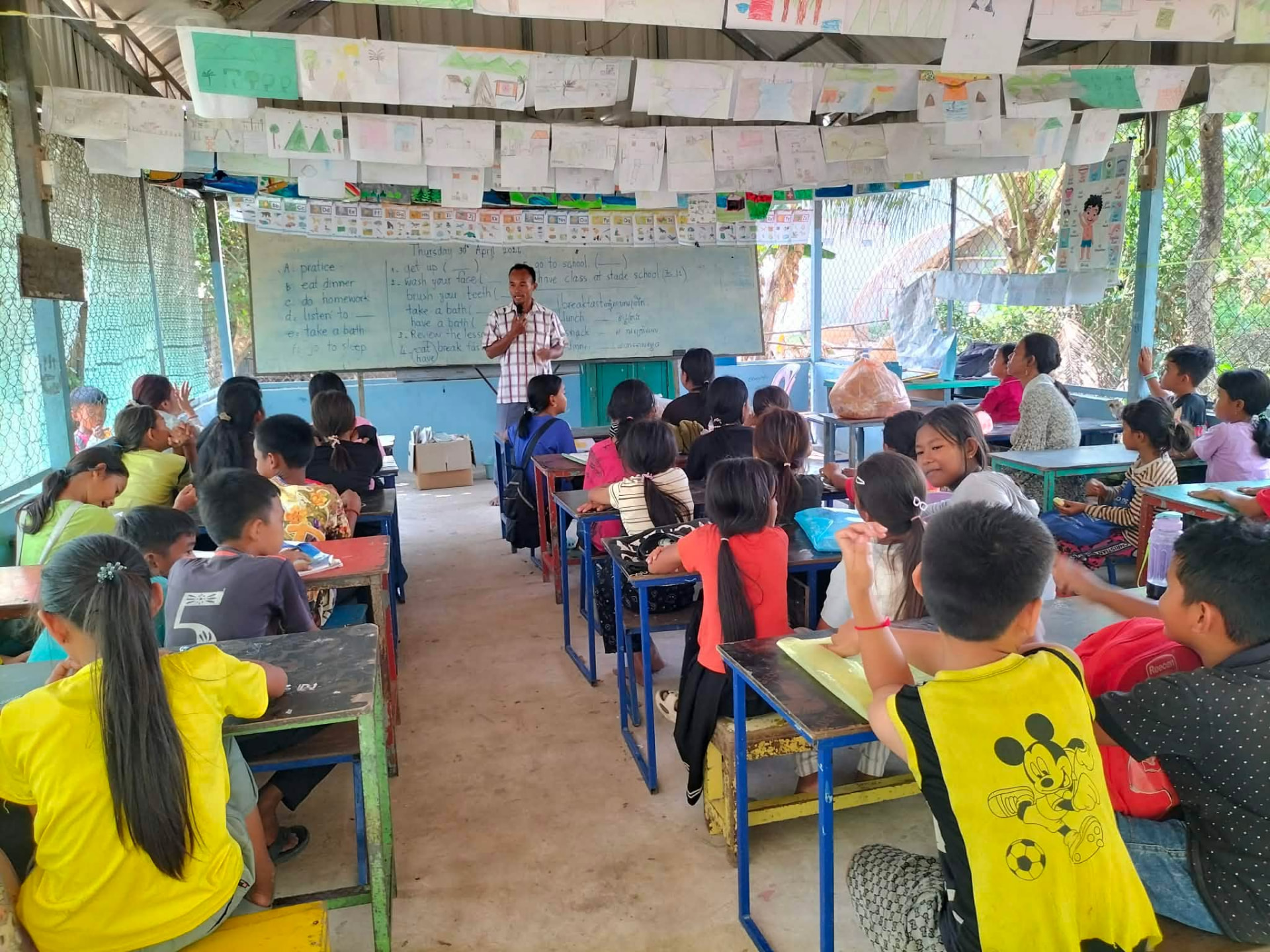 An NFO teacher leading an English lesson in a full village classroom decorated with pupils' drawings